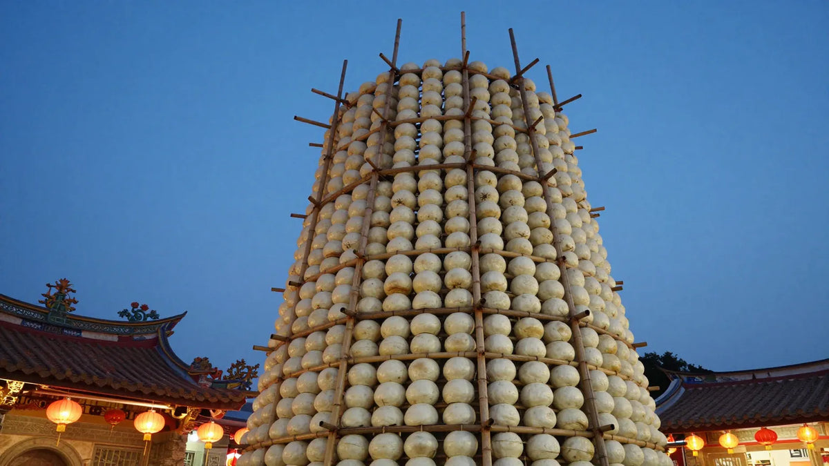 Towering bamboo bun tower covered in white steamed buns at dusk on Cheung Chau island with temple roof and red lanterns
