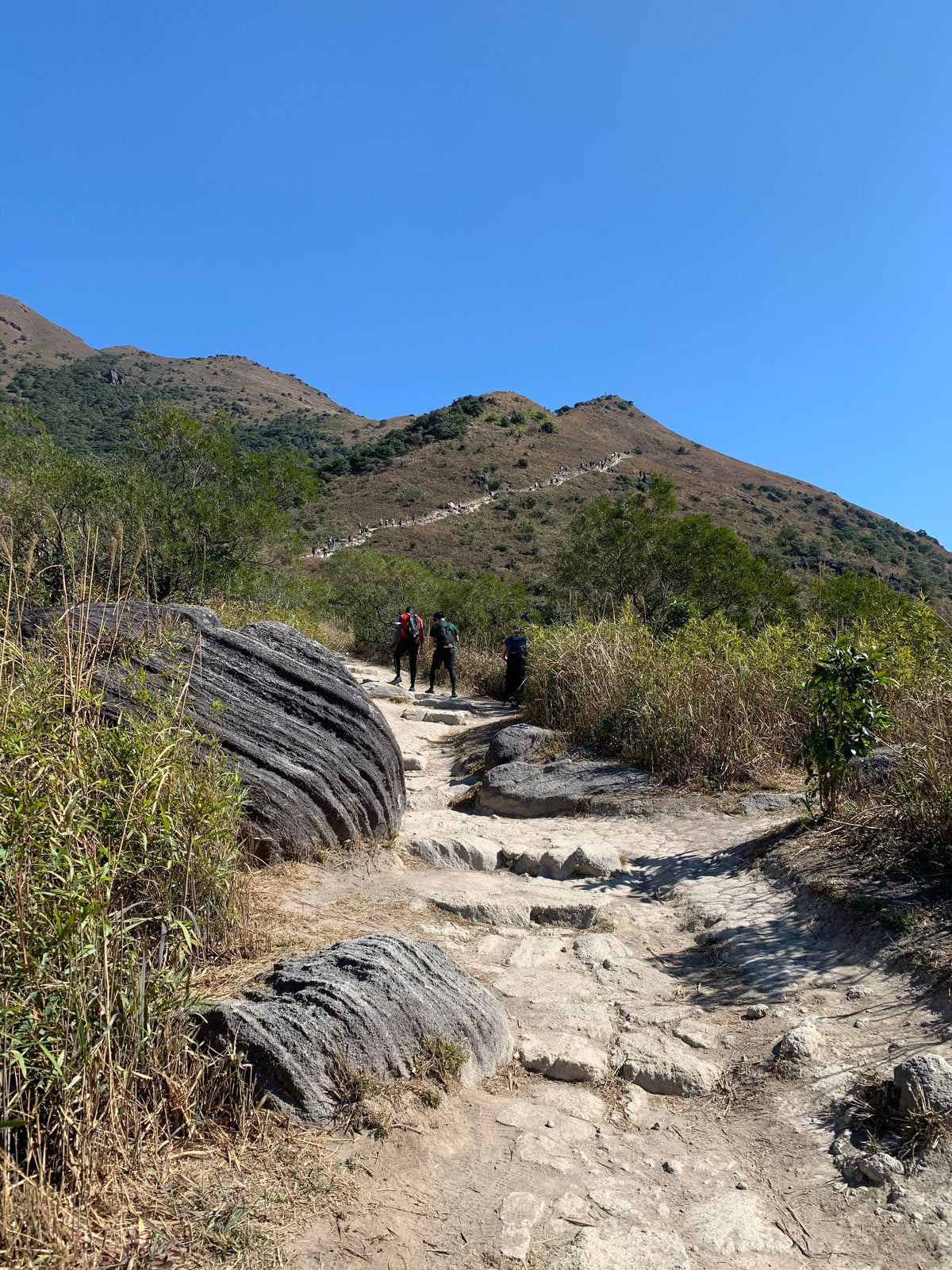 Three hikers exploring a rocky mountain trail in Hong Kong under a clear blue sky