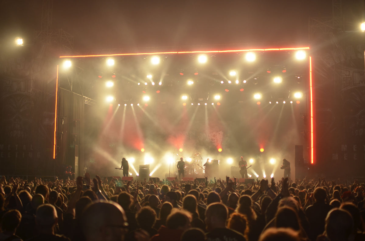 A rock band performing on a concert stage at night with warm brown and tan stage lighting and crowd silhouetted in front